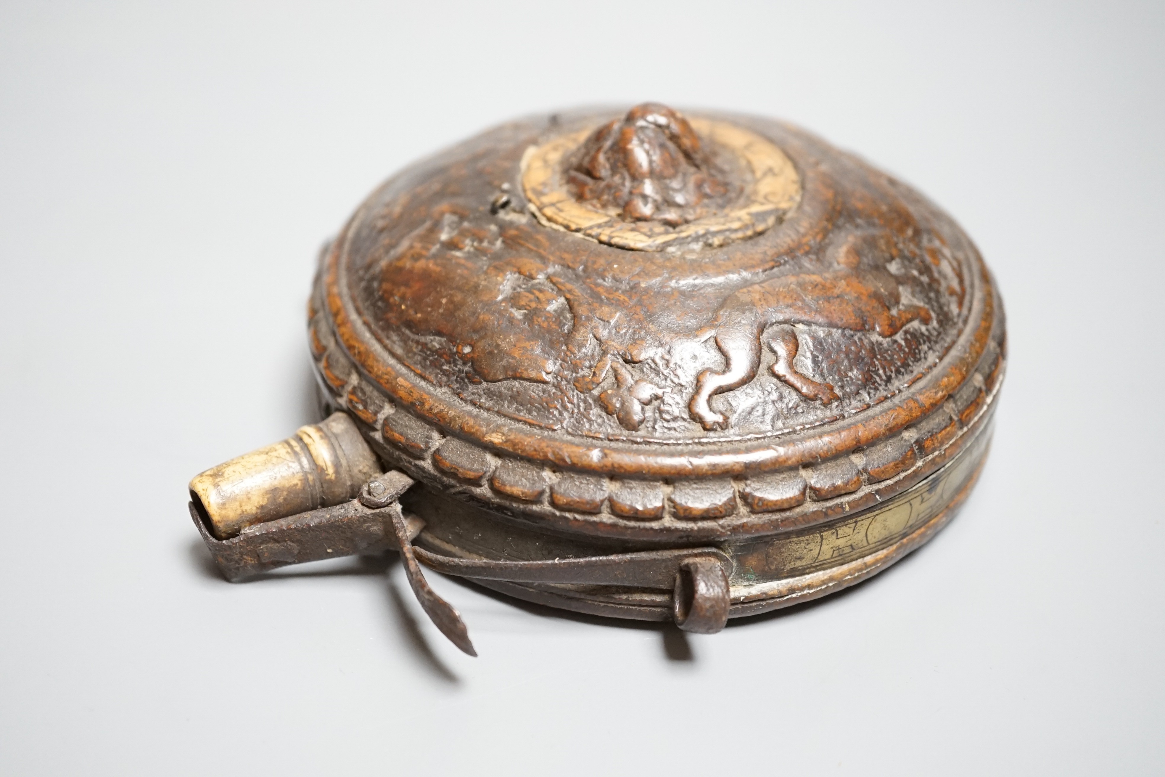 A circular wooden powder flask 17th century, carved with bear, hound and fox, and lion’s mask in low relief surrounded by game counter, engraved brass border, iron lever charger, turned bone nozzle. Diameter 11cms